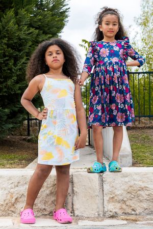 Two children in summer dresses strike confident poses on stone steps, wearing bright pink and turquoise croc sandals.
