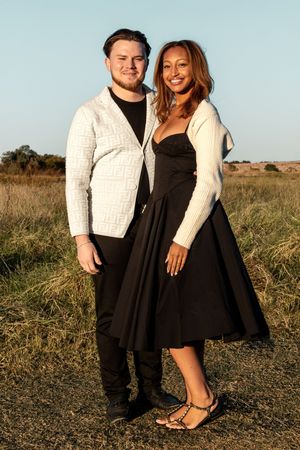 A couple poses together in a grassy field at sunset wearing contrasting black and white outfits.