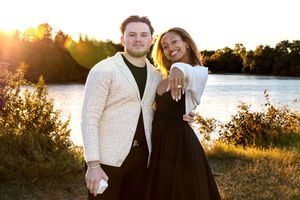 A couple poses together during golden hour sunset by a lake, with warm sunlight filtering through trees.
