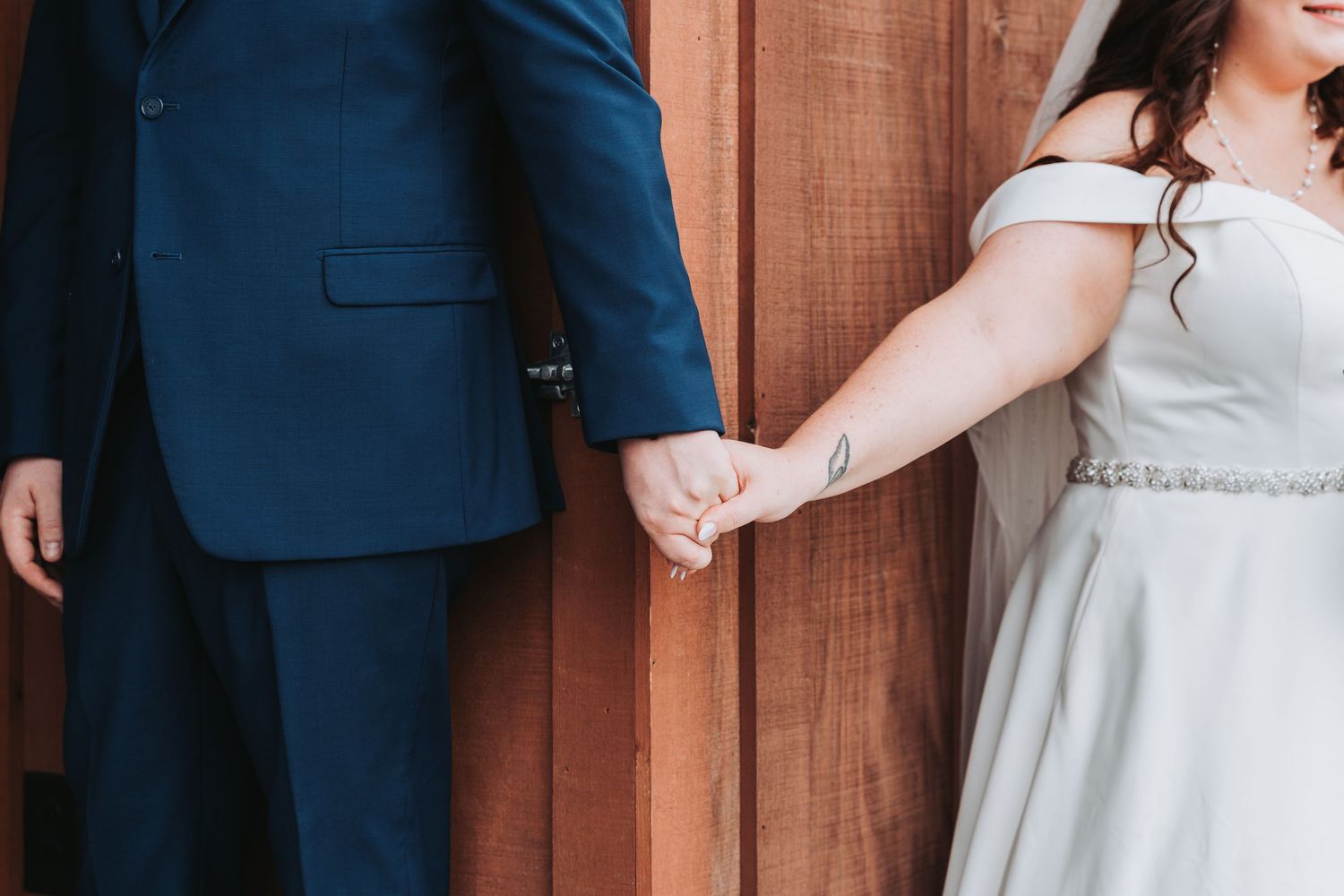 Bride in white dress holds hands with groom in navy suit beside wooden wall during private wedding moment.