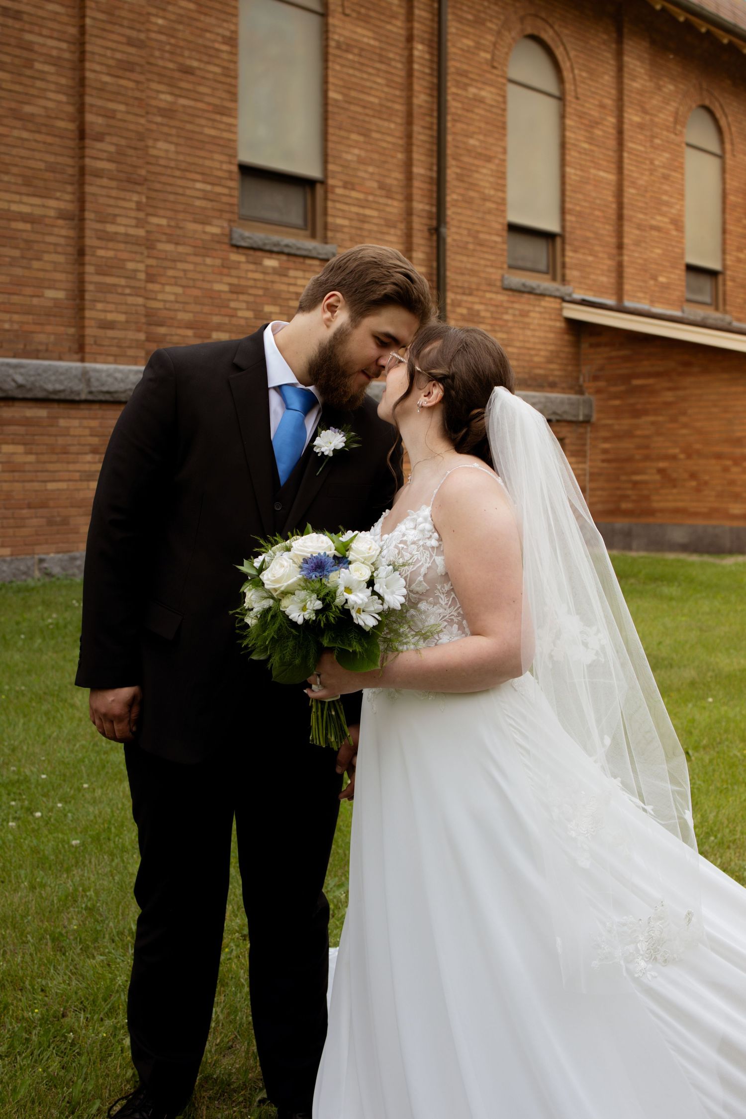 A romantic wedding scene with a bride in white holding flowers beside a groom in black suit and blue tie by a brick church.