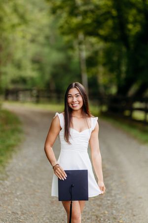 Graduating young woman in white dress holding a black graduation cap on a tree-lined gravel path.