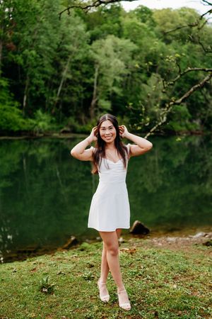 Smiling young woman in white sundress posing by a peaceful forest lake.