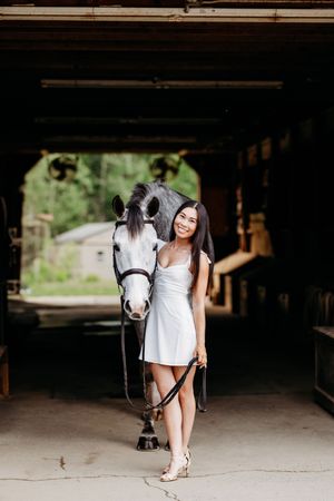 Young woman in white dress smiling while standing with a dark grey horse in a stable.