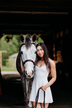 Smiling brunette woman in white dress posing closely with a grey horse in a barn.