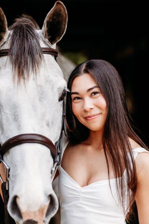 Close-up portrait of a young woman in white dress standing beside a grey horse.