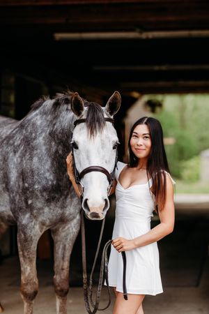 Young woman in white dress standing next to a dapple gray horse with a bridle inside a stable.
