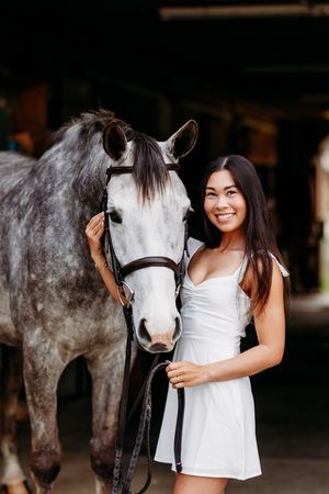 Smiling young woman in white dress posing closely with a dapple gray horse inside a dark stable.