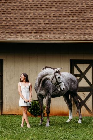 Young woman in white dress laughing beside a dapple gray horse standing on grass near a barn door.