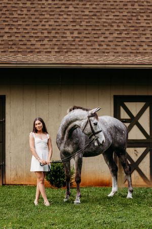 Young woman in white dress standing beside a dapple gray horse on grass in front of a wooden barn.