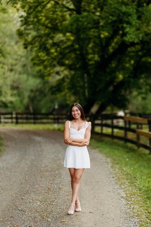 Young woman in white mini dress standing on a tree-lined gravel path with arms crossed, smiling.