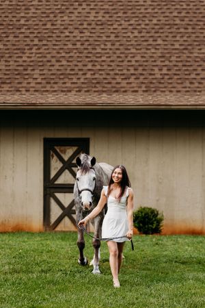 Young woman in white dress smiling while leading a dapple gray horse across a green lawn by a barn.