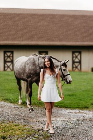 Smiling young woman in white dress walking on gravel path with a dapple gray horse near a barn.