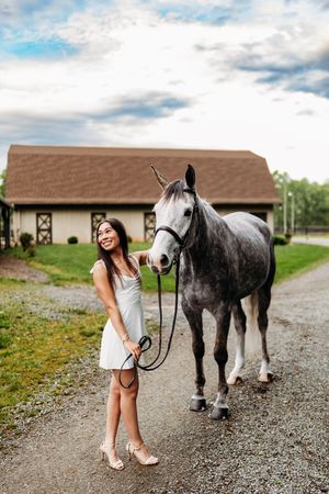 Young woman in white dress smiling while leading a dapple gray horse on a gravel path near a barn.
