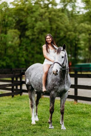 Smiling young woman in white dress riding a dapple gray horse bareback in a green paddock.