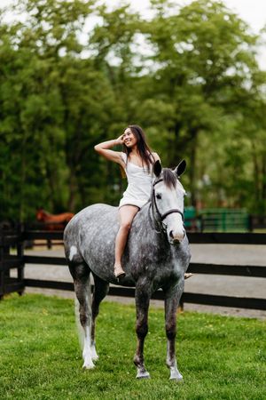 Young woman in white dress sitting bareback on a dapple gray horse in a green paddock.
