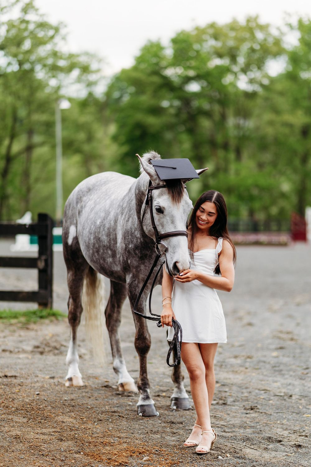 Smiling graduate in white dress with dapple gray horse wearing a graduation cap at stable.
