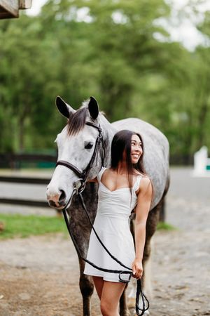 Cheerful young woman in white dress leading a dapple gray horse on a gravel stable path.