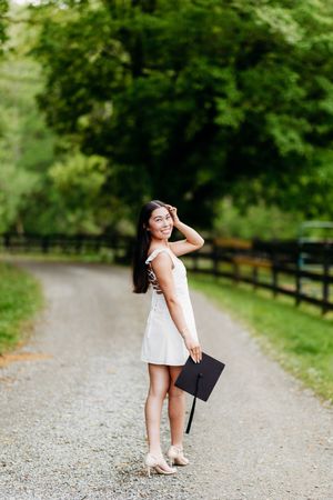 Smiling graduate in white dress holding mortarboard cap on a scenic tree-lined gravel path.