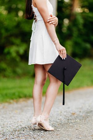 Close-up of graduate in white dress and heels holding a black mortarboard cap outdoors.