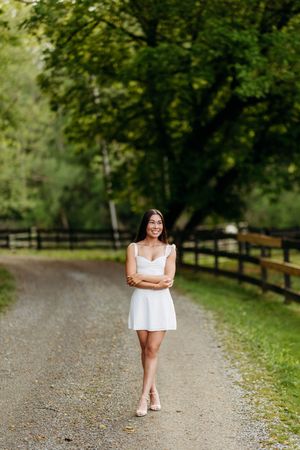 Happy young woman in white dress walking on a lush tree-lined gravel road with crossed arms.