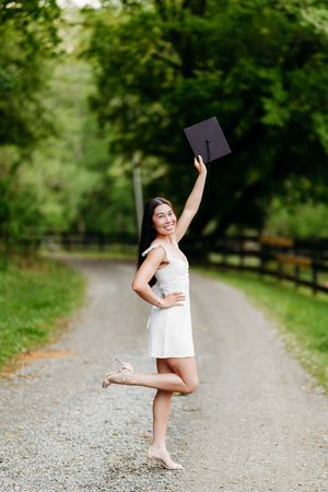 Excited graduate in white dress raising mortarboard cap while balancing on one leg outdoors.