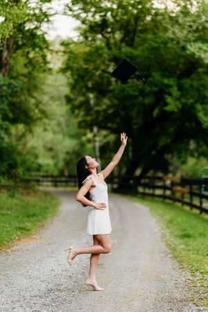 Joyful graduate in white dress tossing mortarboard cap in the air on a tree-lined path.