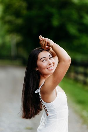 Smiling young woman with long dark hair posing in white dress on a green tree-lined path.