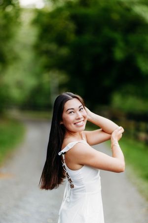 Happy young woman in white sundress posing with hands raised on a lush green outdoor path.