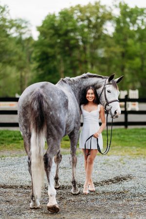 Smiling woman in white dress leading grey horse on gravel path.