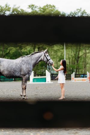 Young woman in white dress standing beside a dapple gray horse at an equestrian arena.