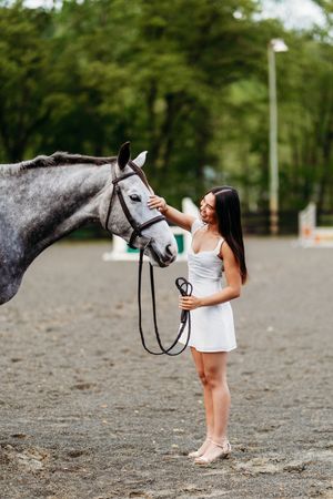 Young woman in white dress petting grey horse in sandy arena.