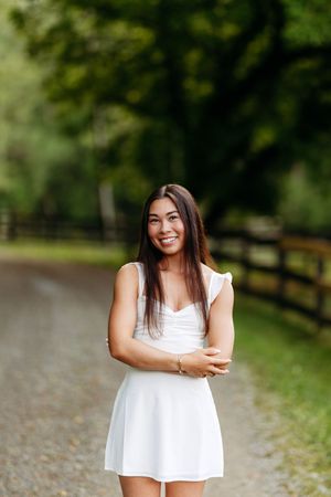 Smiling young woman in white sleeveless dress posing on a scenic gravel path bordered by wooden fences.