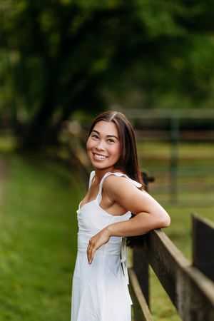 Smiling young woman in white dress leaning on a wooden fence in a lush green field.