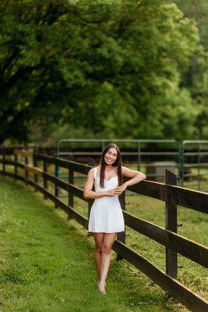 Young woman in white sundress smiling while leaning on a wooden fence in a green pasture.