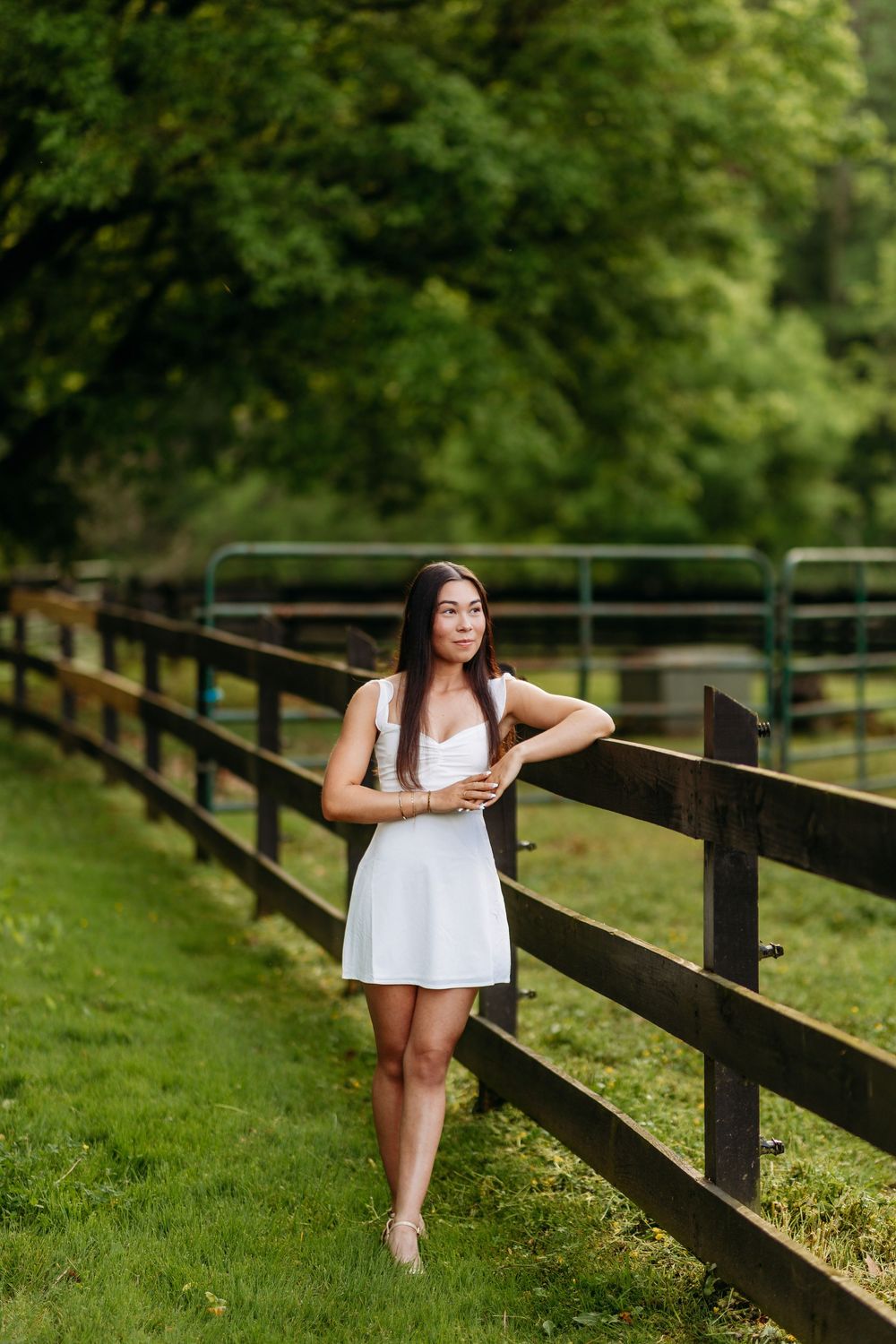 Young woman in white dress leaning against a wooden farm fence surrounded by greenery.