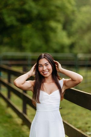 Smiling young woman in white dress posing with hands in hair by a wooden fence.