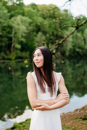 Young woman in white ruffled dress standing by a serene green lake in nature.