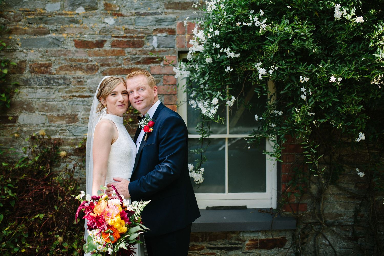 A couple embraces near a rustic stone wall with blooming flowers during a wedding photoshoot.