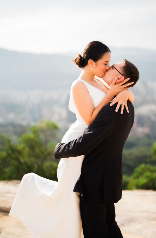 A romantic portrait shows embrace between couple in wedding attire with city vista background during sunset.