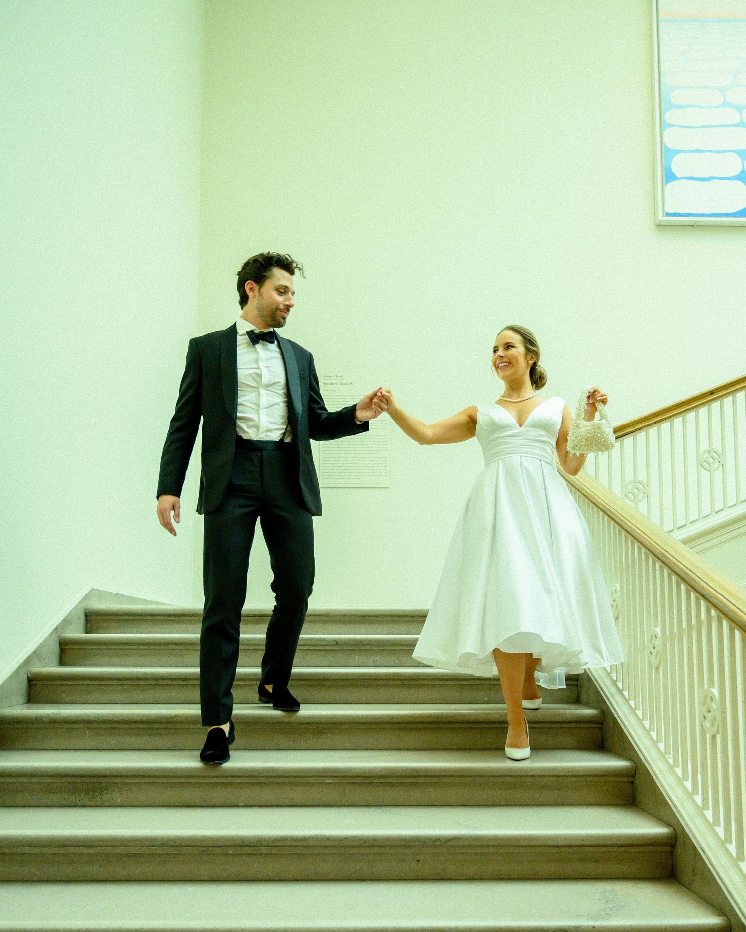 A couple in formal wedding attire hold hands while descending a staircase in a mint green hallway.