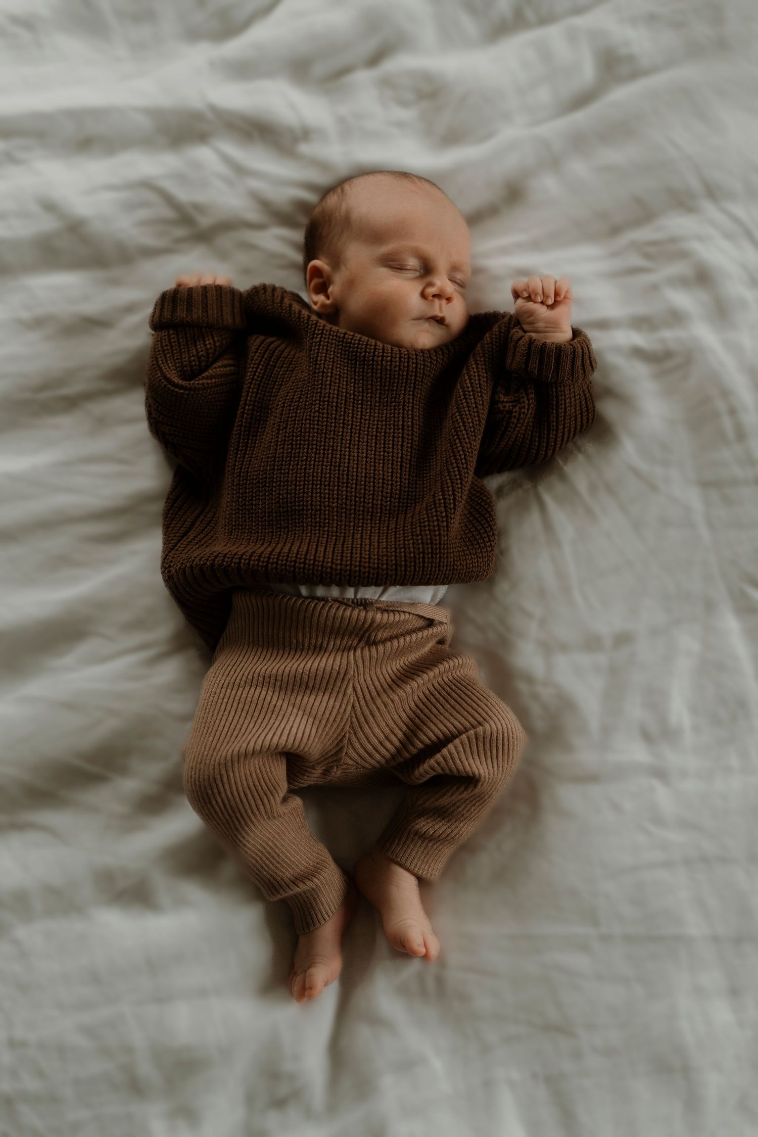 A sleeping baby wearing a brown sweater and tan ribbed pants lies peacefully on white bedding.