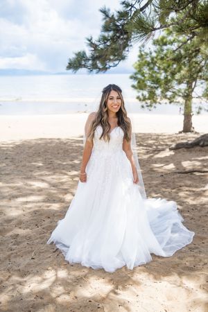 A flowing white wedding dress flowing in the breeze on a sandy beach with pine trees in the background.