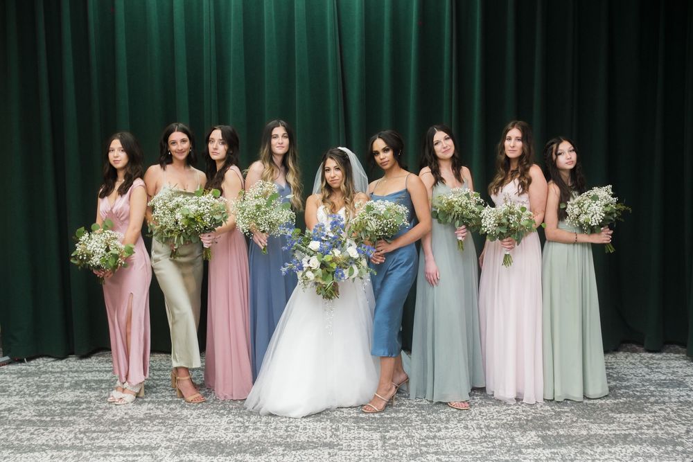 A bridal party poses together in pastel-colored dresses holding white and green floral bouquets against a dark green backdrop.