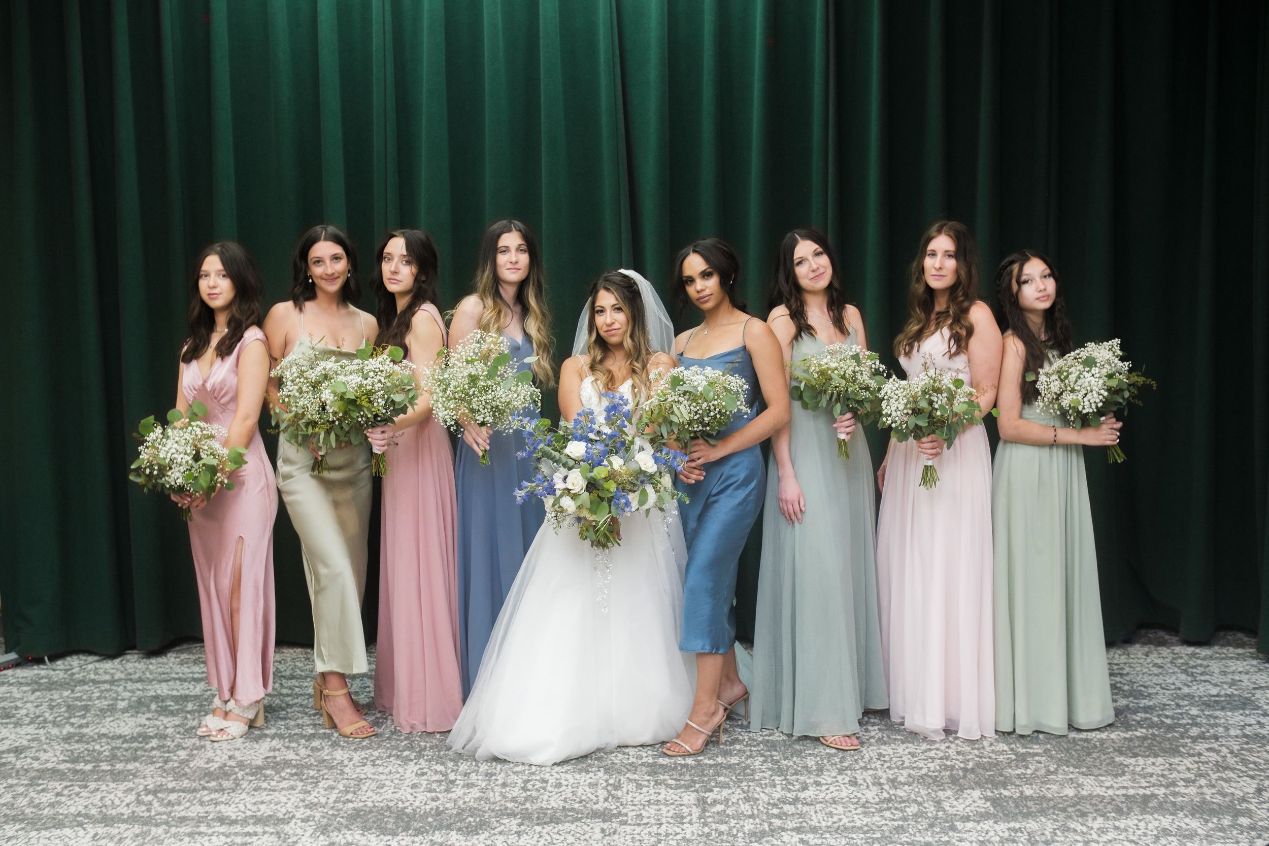 A bridal party poses together in pastel-colored dresses holding white and green floral bouquets against a dark green backdrop.