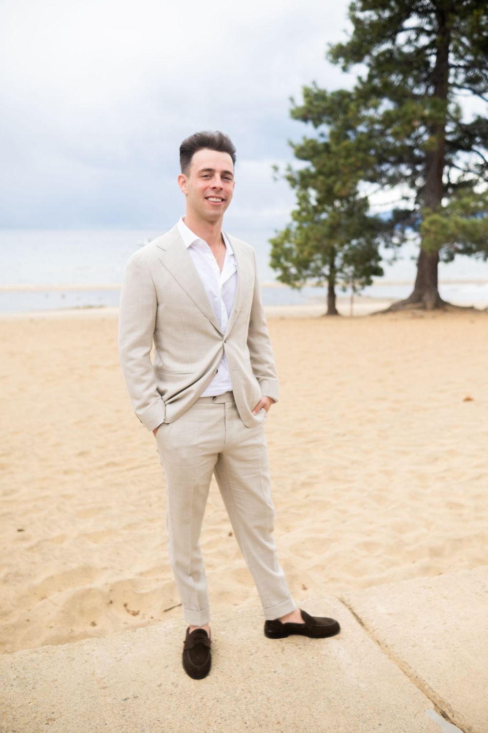 Someone wearing a light beige suit and white shirt poses playfully on a sandy beach with pine trees in the background.