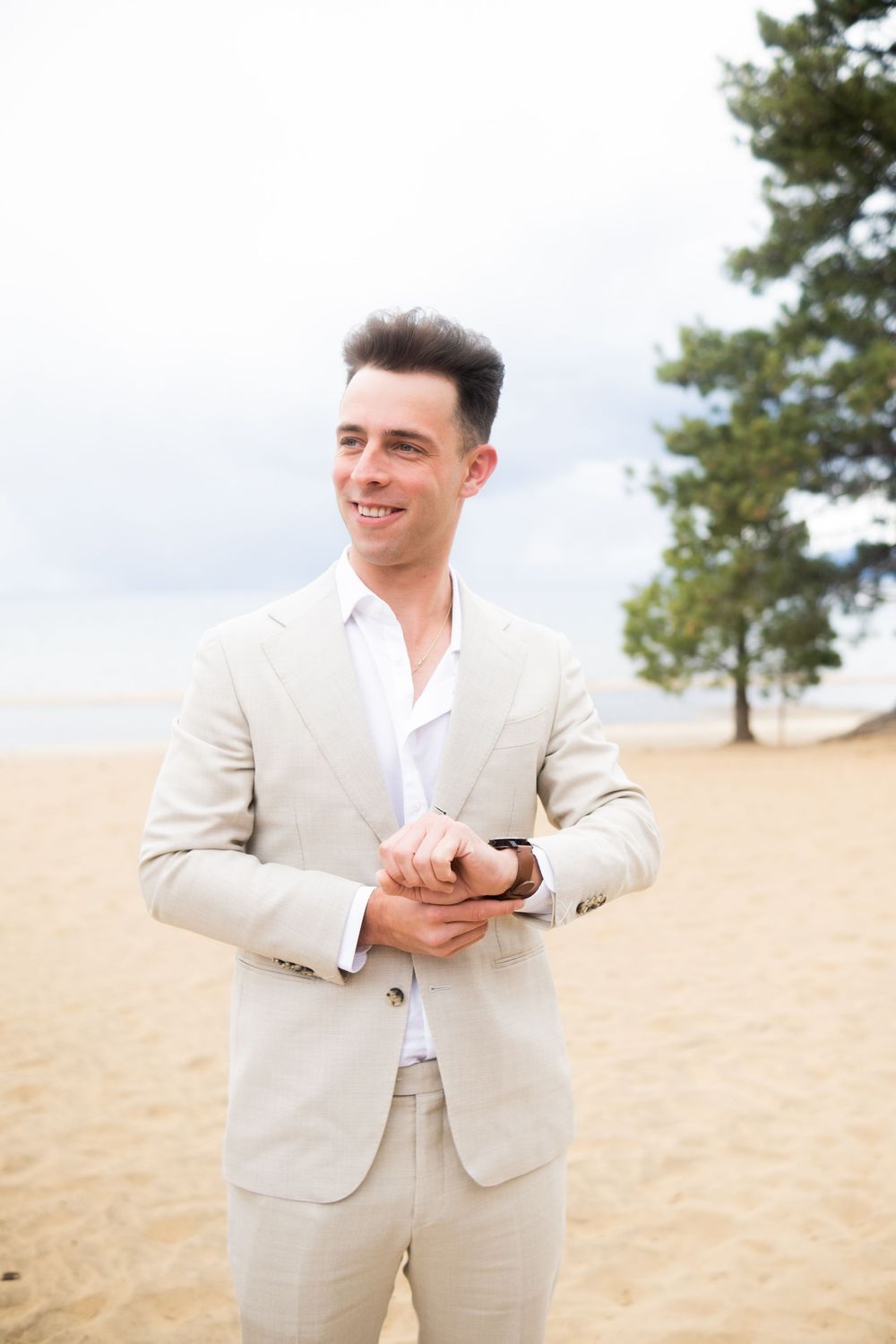 Person in light beige suit smiles while checking watch on sandy beach with pine trees in background.