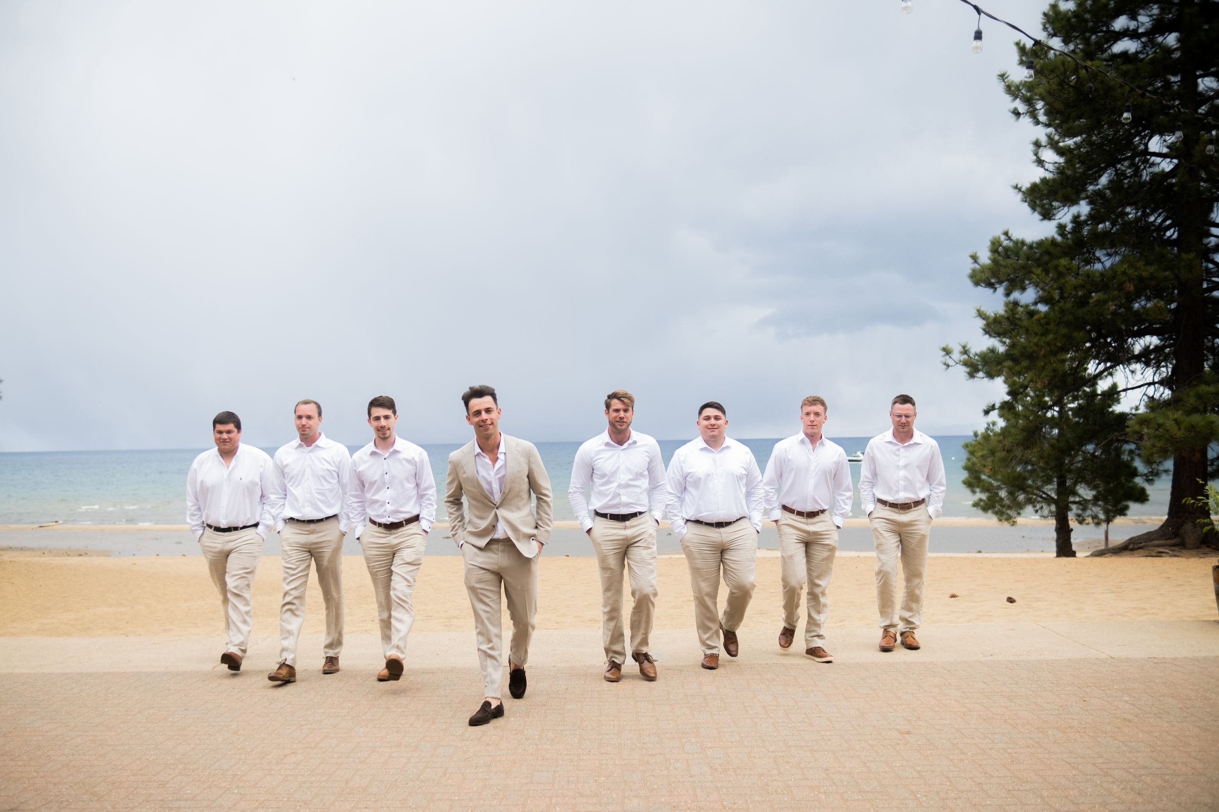 Groomsmen in matching white shirts and khaki pants walk in line formation on a scenic beach with cloudy skies.