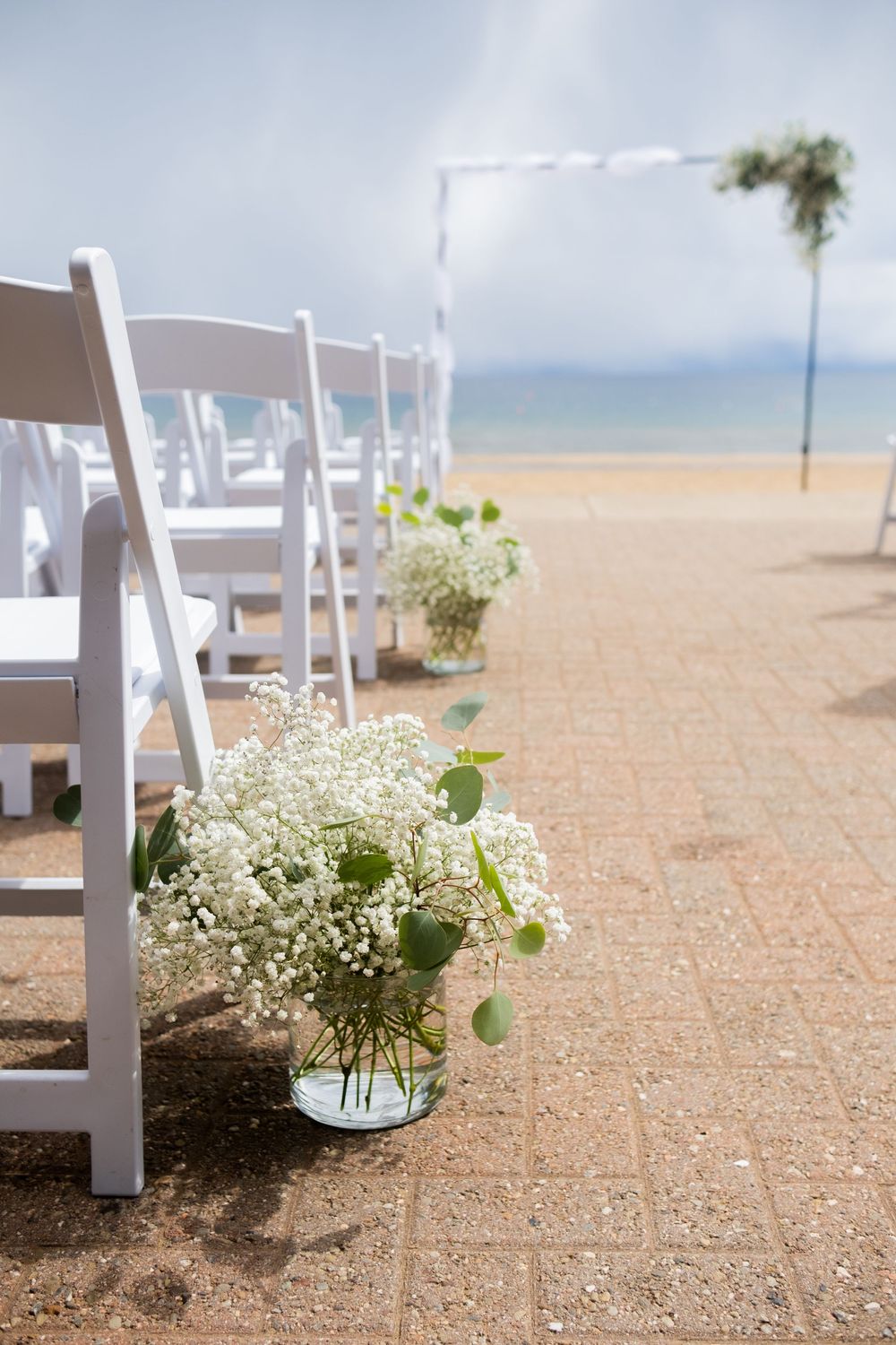White folding chairs and delicate baby's breath floral arrangements line an aisle at a beachfront wedding ceremony setup.