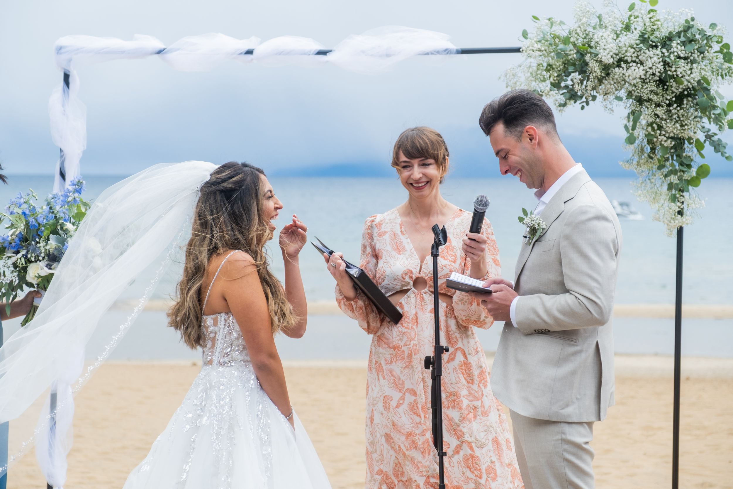 Outdoor beach wedding ceremony with three people standing together under floral arch by the ocean.